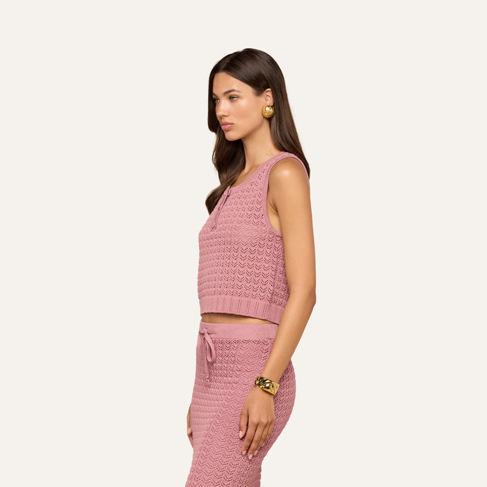 A woman with long brown hair stands in profile against a plain, light-colored background, wearing the Carven Pointelle Knit Tank in pink as part of a matching sleeveless top and skirt set. She accessorizes with gold earrings and a gold bracelet.