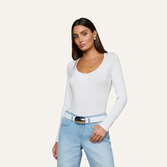 A woman wearing the Cassandra Ruched Bodysuit with long sleeves and a scoop neckline, paired with light blue jeans and a white belt, stands against a plain light background with one hand resting on her hip.