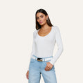 A woman wearing the Cassandra Ruched Bodysuit with long sleeves and a scoop neckline, paired with light blue jeans and a white belt, stands against a plain light background with one hand resting on her hip.
