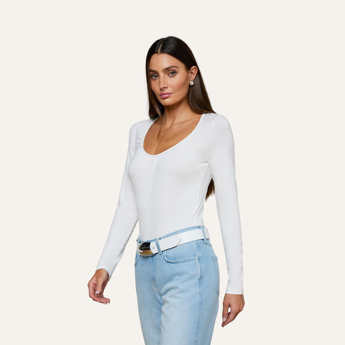Woman with long brown hair wearing the Cassandra Ruched Bodysuit—a white long sleeve top with a scoop neckline—paired with light blue jeans and a white belt, standing against a plain light background.