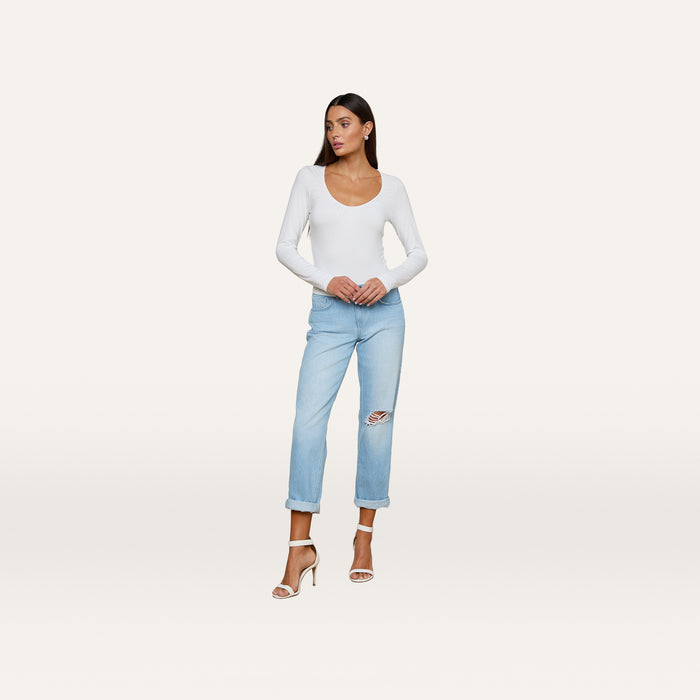 The woman is wearing the Cassandra Ruched Bodysuit, a white long-sleeve top with a scoop neckline, paired with light blue ripped jeans and high-heeled sandals. She stands against a plain light background, looking to the side with her hands resting on her jeans.