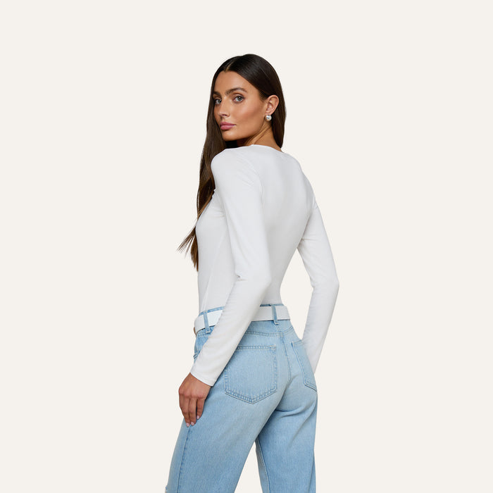 A woman with long brown hair, wearing the Cassandra Ruched Bodysuit—a white long sleeve top with a scoop neckline—paired with light blue jeans, stands sideways and looks over her shoulder against a plain, light background.