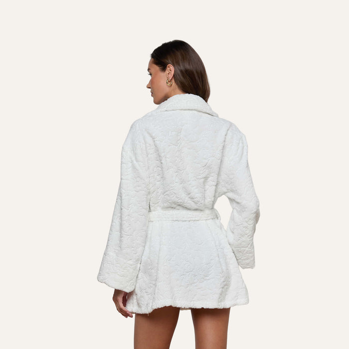 A woman with long brown hair stands facing away from the camera against a plain light background, wearing the Caterina Cover-Up Robe—a white, textured cotton kimono robe with a belt.