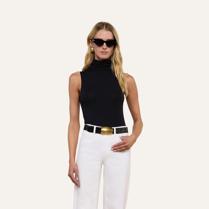 A woman wearing the Ceci Sleeveless Turtleneck in a slim fit, paired with white pants featuring a large gold buckle belt and black sunglasses, stands against a plain light background.