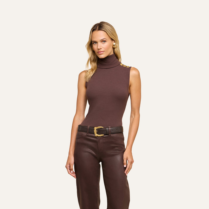 A woman stands against a plain light background wearing a slim-fit Ceci Sleeveless Turtleneck in brown, featuring gold buttons on the shoulder, paired with dark brown pants, a black belt with a gold buckle, and gold earrings.