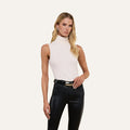 A woman with long blonde hair poses against a plain, light background, wearing the Ceci Sleeveless Turtleneck in white—its slim fit accentuating her silhouette—as well as black pants, with one hand placed on her waist.