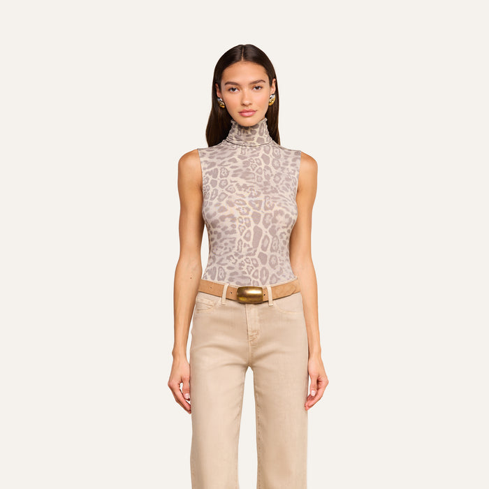 A woman stands facing forward, wearing the Ceci Sleeveless Turtleneck in a grey leopard print, paired with beige pants, a wide belt with a gold buckle, and small hoop earrings. She has long straight hair and a neutral expression.