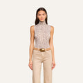 A woman stands facing forward, wearing the Ceci Sleeveless Turtleneck in a grey leopard print, paired with beige pants, a wide belt with a gold buckle, and small hoop earrings. She has long straight hair and a neutral expression.