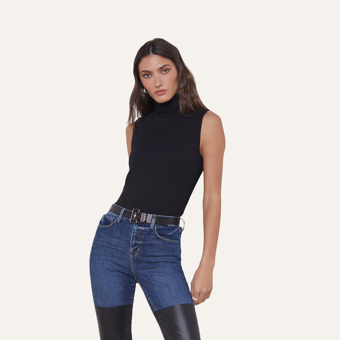 A woman wearing the Ceci Sleeveless Turtleneck and blue jeans with black leather patches, standing against a plain white background.