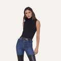 A woman wearing the Ceci Sleeveless Turtleneck and blue jeans with black leather patches, standing against a plain white background.
