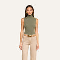 A woman models the Ceci Sleeveless Turtleneck in ash olive green, styled with beige high-waisted pants, a wide brown belt, and gold earrings, standing against a plain light background.