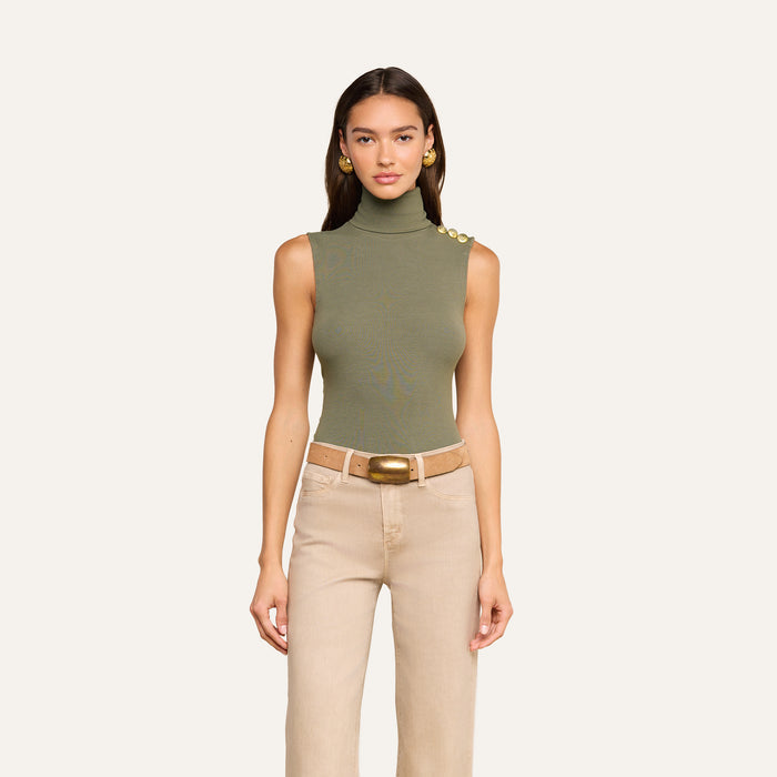 A woman models the Ceci Sleeveless Turtleneck in ash olive green, styled with beige high-waisted pants, a wide brown belt, and gold earrings, standing against a plain light background.