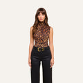 A woman stands against a plain background in the Ceci Sleeveless Turtleneck, which she has tucked into slim-fit black pants secured with a wide belt featuring a large gold buckle. She wears her long brown hair down and has a neutral expression.