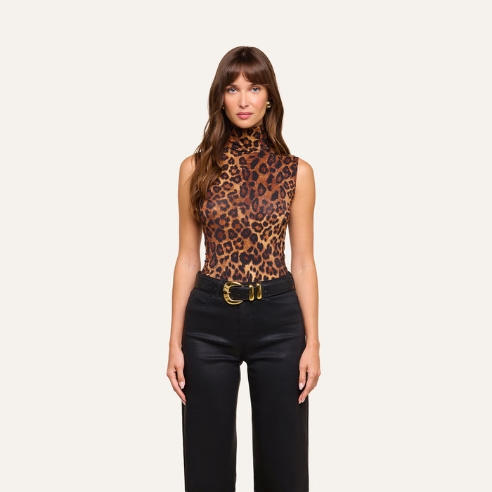 A woman stands against a plain background in the Ceci Sleeveless Turtleneck, which she has tucked into slim-fit black pants secured with a wide belt featuring a large gold buckle. She wears her long brown hair down and has a neutral expression.