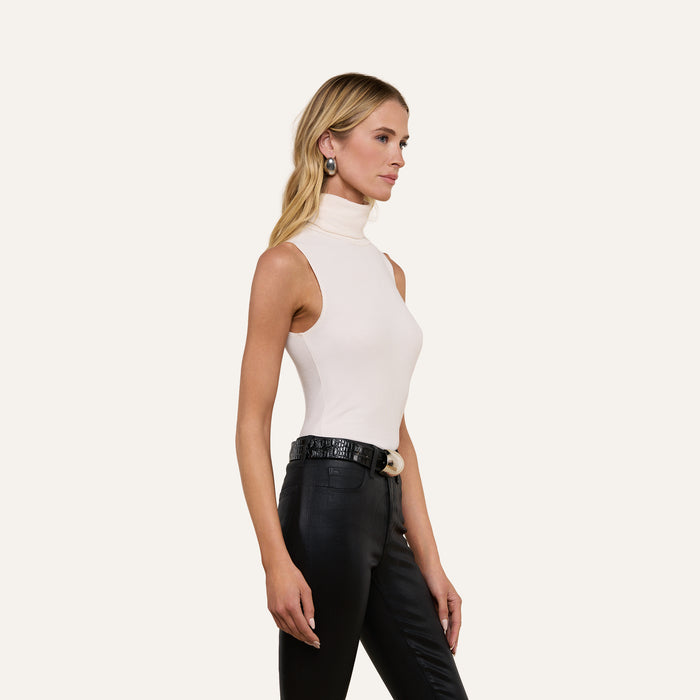 A woman with long blonde hair and earrings stands against an off-white background, facing right, wearing the Ceci Sleeveless Turtleneck in white paired with black high-waisted pants.