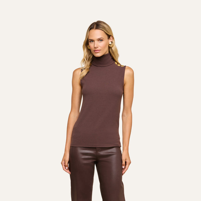 A woman stands against a plain background wearing the Ceci Sleeveless Turtleneck—a slim-fit, sleeveless brown top with gold buttons on the shoulder—paired with matching brown pants. Made from stretch rib knit, the turtleneck complements her long, wavy blonde hair as she looks directly at the camera.