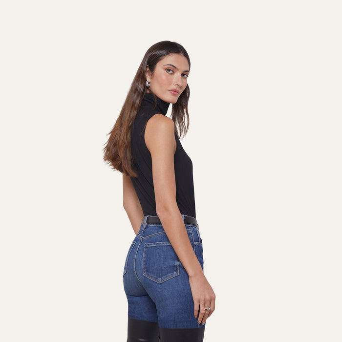 Woman with long brown hair wearing the Ceci Sleeveless Turtleneck—a slim-fit black top—paired with high-waisted two-tone blue jeans, standing sideways and looking toward the camera against a plain light background.