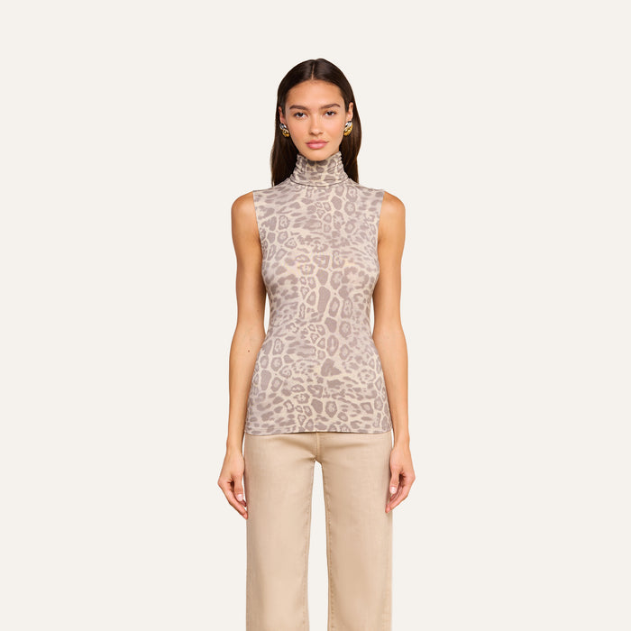 A woman stands facing forward against a plain off-white background, wearing the Ceci Sleeveless Turtleneck in a slim fit grey leopard print paired with light-colored pants.
