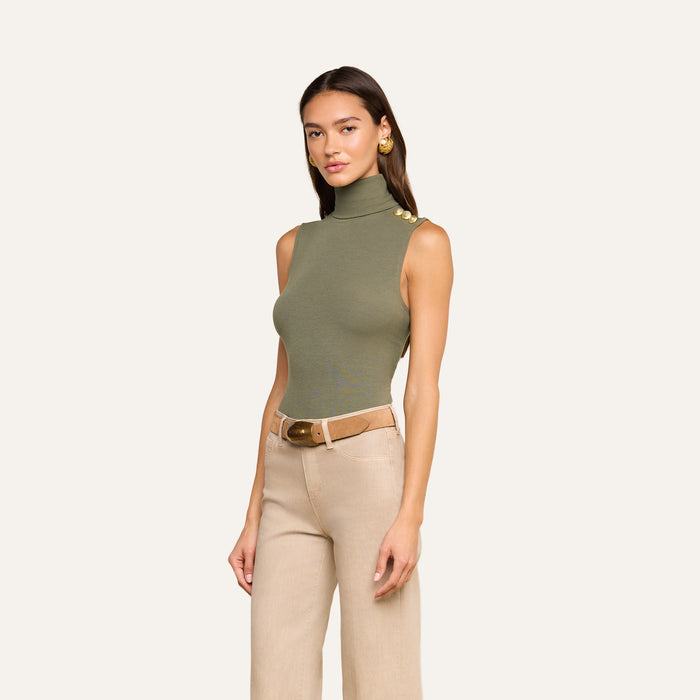 A woman stands against a plain light background wearing the Ceci Sleeveless Turtleneck in ribbed knit ash olive green with gold shoulder buttons, paired with beige pants and a brown belt.