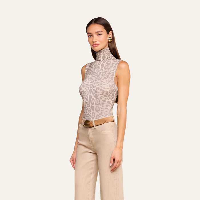 Wearing the Ceci Sleeveless Turtleneck in a slim fit grey leopard print, paired with beige pants and a brown belt, a woman stands against a plain light background.