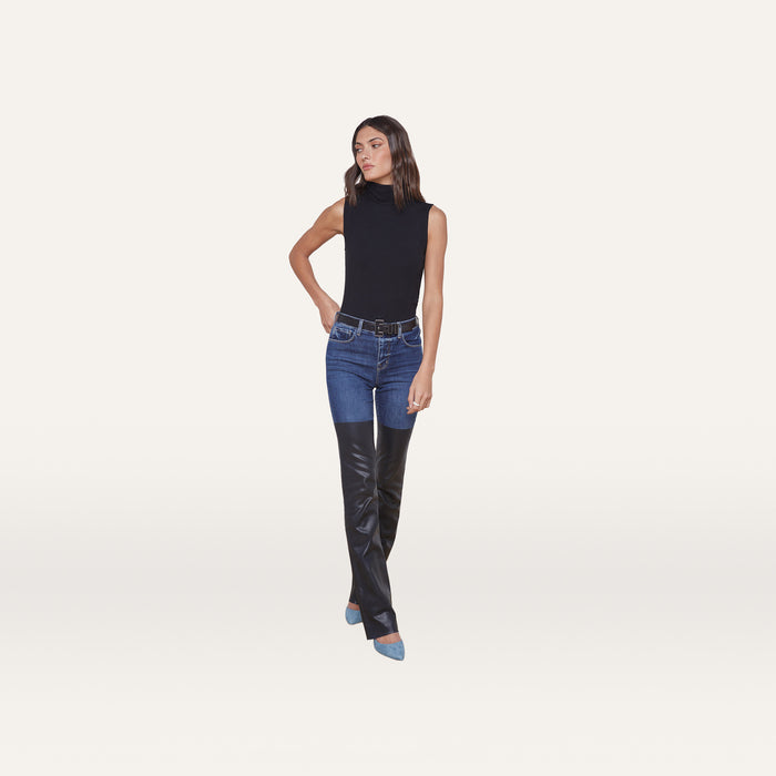 A woman wearing the Ceci Sleeveless Turtleneck, blue jeans with black leather panels on the lower legs, and pointed blue heels stands against a plain, light background.