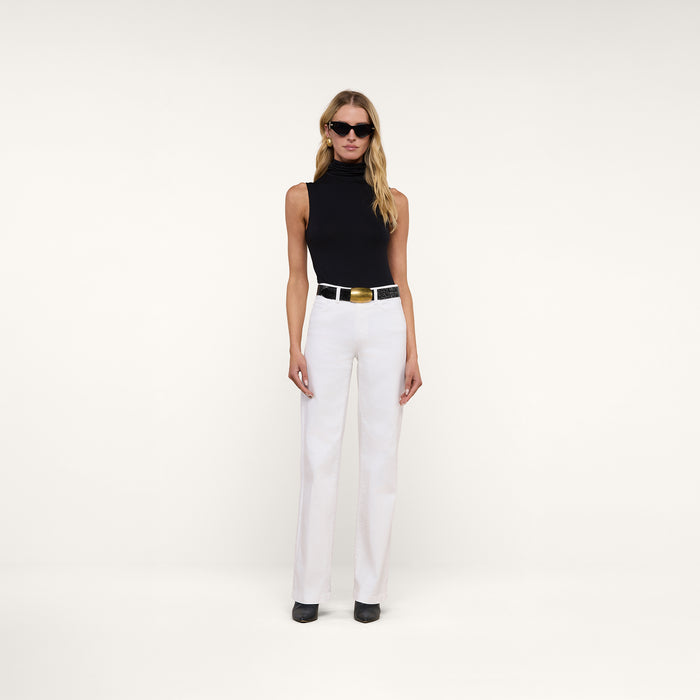 A woman wearing sunglasses, the Ceci Sleeveless Turtleneck in black, white pants with a wide belt, and black shoes stands against a plain white background.