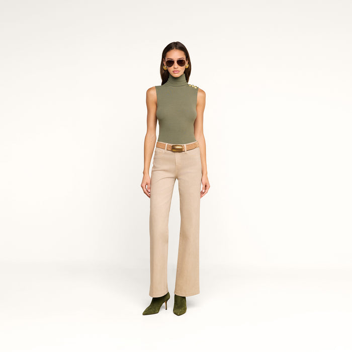 Wearing the Ceci Sleeveless Turtleneck in ash olive green, paired with beige high-waisted belted pants and green heels, a woman in sunglasses stands against a plain white background.