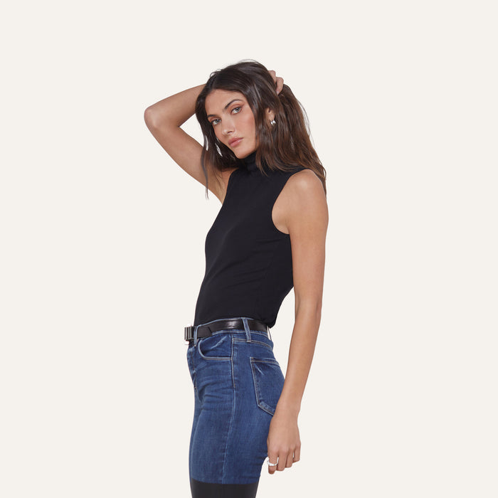 Woman in a slim-fit Ceci Sleeveless Turtleneck and blue jeans stands sideways, looking at the camera with one hand resting on her head against a plain white background.