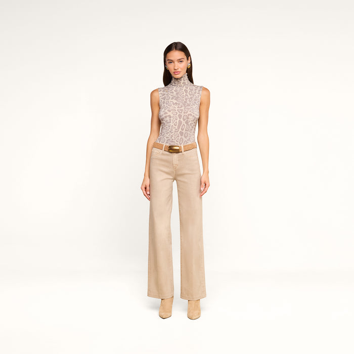 A woman with straight brown hair and a neutral expression stands against a white background, wearing the Ceci Sleeveless Turtleneck in a grey leopard print, paired with beige wide-leg pants featuring a belt and matching heeled boots.