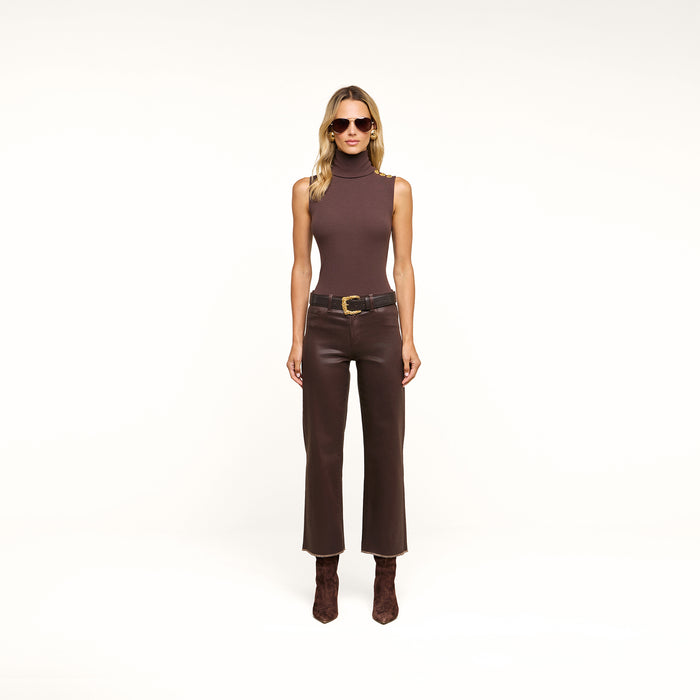 A woman wearing sunglasses, a Ceci Sleeveless Turtleneck slim-fit stretch rib knit top, brown leather pants, a black belt, and brown boots stands against a plain white background.