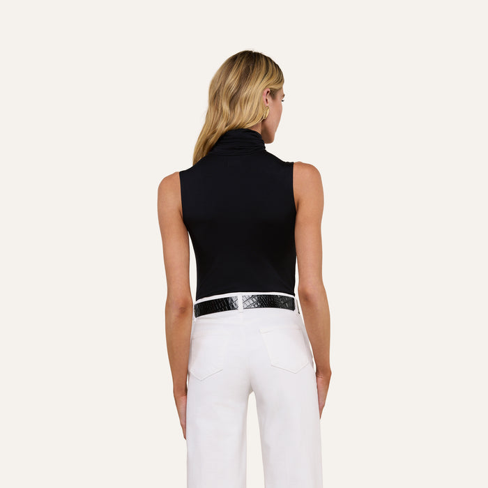 A woman with long blonde hair is shown from the back, wearing the Ceci Sleeveless Turtleneck and white pants with a black textured belt, standing against a plain light background.