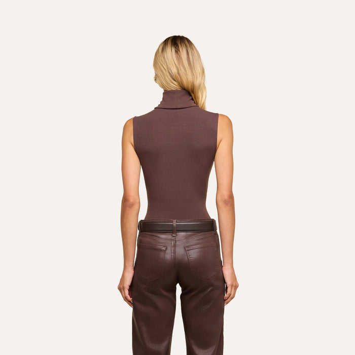 A woman with blonde hair stands with her back to the viewer against a plain light background, wearing a slim-fit, sleeveless brown Ceci Sleeveless Turtleneck and brown leather pants.