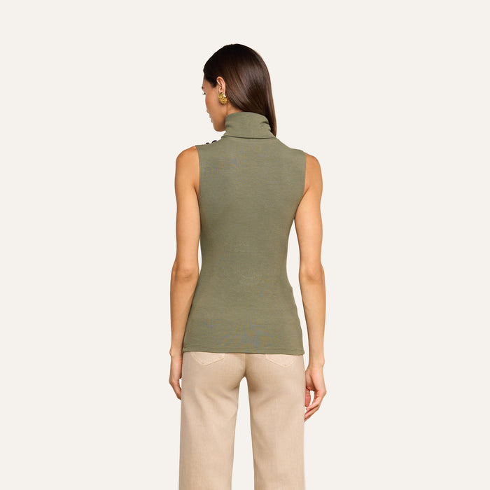 A woman stands facing away, wearing the Ceci Sleeveless Turtleneck in ash olive green and beige pants against a plain light background.