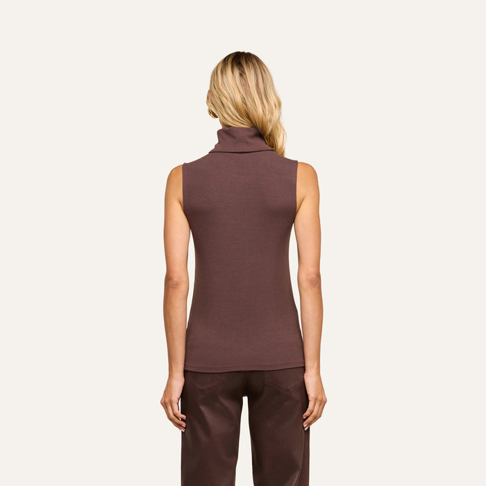 A person with long blonde hair, wearing the Ceci Sleeveless Turtleneck in a slim-fit brown rib knit and matching brown pants, is standing facing away against a plain light background.