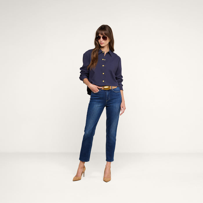 A woman stands against a plain white background with one hand in her pocket, wearing sunglasses, a navy button-up shirt, tan high heels, and the Celestia Slim-Leg Jean in a vintage wash with a relaxed fit.