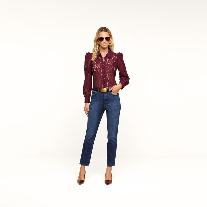 A woman wearing sunglasses, a maroon lace long-sleeve top, the Celestia Slim-Leg Jean in a dark indigo wash, and maroon heels poses against a plain white background with one hand in her pocket.