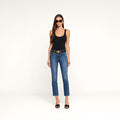 A woman stands against a plain white background wearing black sunglasses, a black tank top, the Celestia Slim-Leg Jean with a belt, and black heels.
