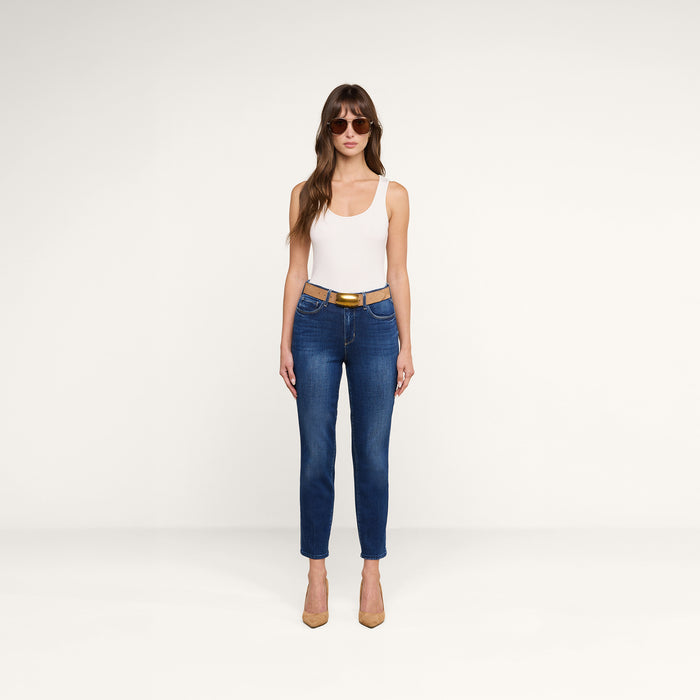 A woman wearing sunglasses, a white tank top, the Celestia Slim-Leg Jean in a vintage wash with a tan belt, and tan heels stands facing forward against a plain white background.