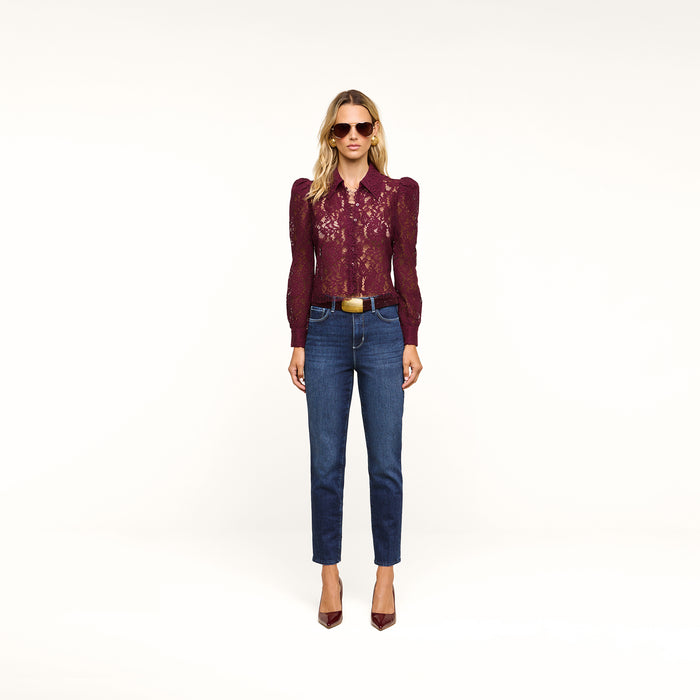 A woman stands against a plain background wearing sunglasses, a burgundy lace blouse, and the Celestia Slim-Leg Jean in classic stretch denim with an ultra high rise fit. She completes her look with a brown belt and burgundy heels.