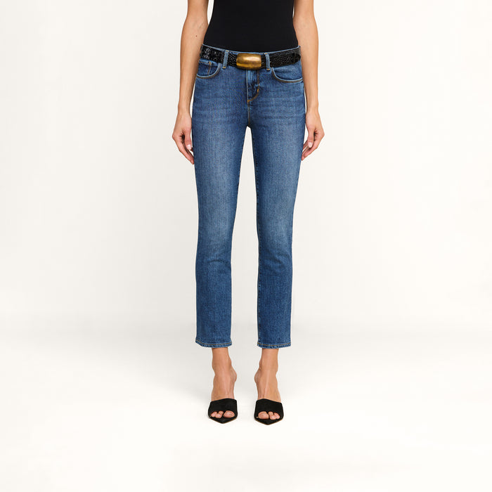 Person wearing Celestia Slim-Leg Jean in ultra high rise blue stretch denim, a black belt with a large buckle, a black top, and black open-toe shoes, standing against a plain white background.