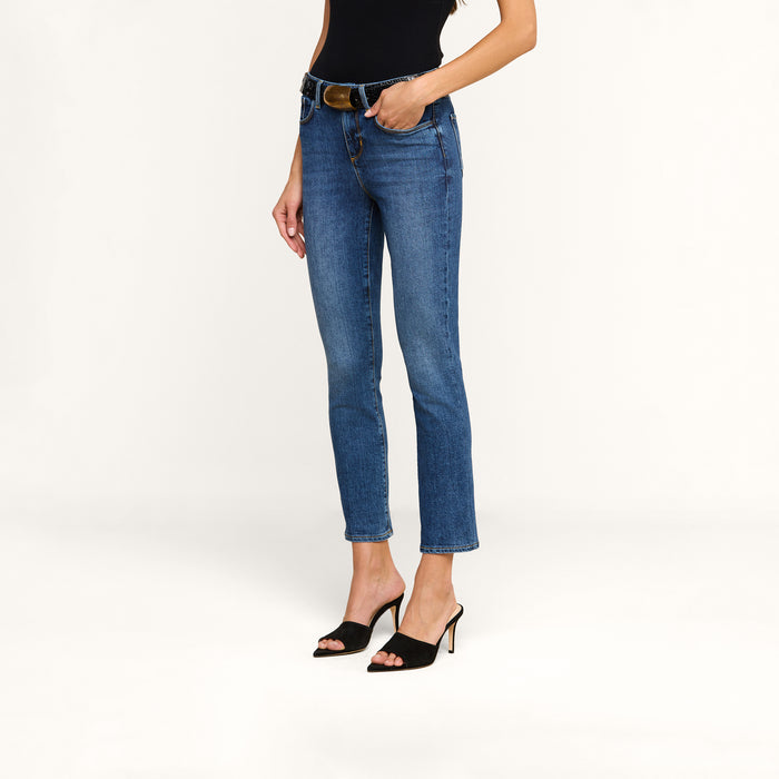 A woman wearing the Celestia Slim-Leg Jean in ultra high rise blue stretch denim, paired with a black sleeveless top, black belt, and black open-toe heeled sandals, stands against a plain white background. Her head is not visible in the image.