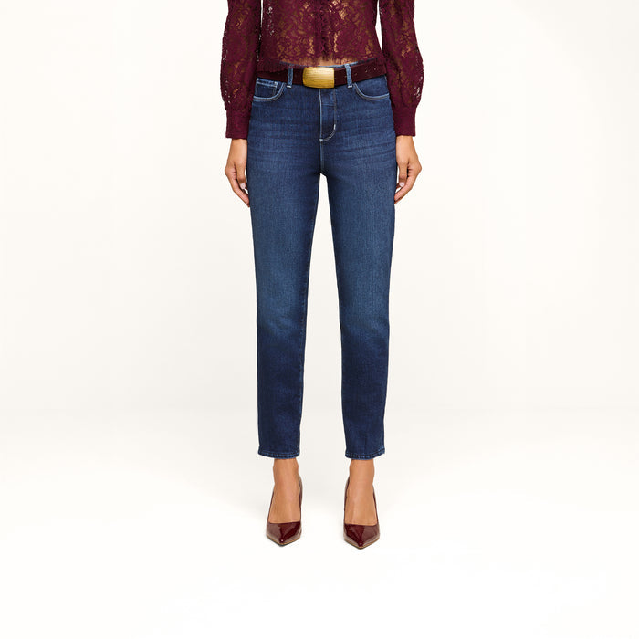 A woman models the Celestia Slim-Leg Jean in a classic dark indigo wash, styled with a sheer burgundy lace top and matching pointed heels. The photo is cropped to show only her lower body and torso against a plain white background.