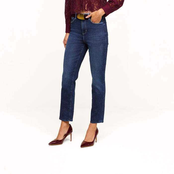 A woman stands against a white background, shown from the shoulders down, wearing the Celestia Slim-Leg Jean in a dark indigo wash with classic stretch denim, paired with a burgundy lace top and matching burgundy high heels.