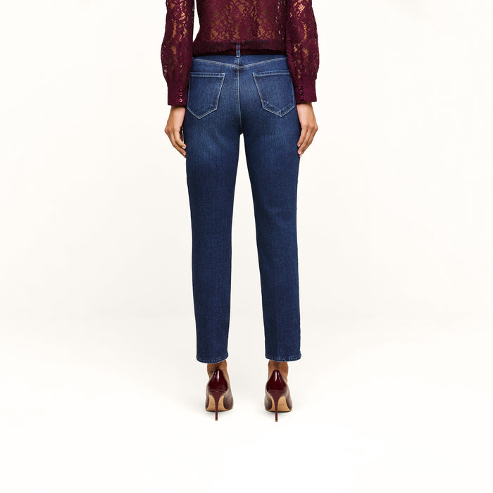 A person stands with their back to the camera, wearing a burgundy long-sleeve lace top, Celestia Slim-Leg Jean in a dark indigo wash stretch denim, and burgundy high-heeled shoes against a plain white background.