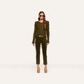 A woman wears the Celestia Velvet Slim-Leg Jean in dark green, paired with an olive green sweater, a brown belt, and beige heels. She has long wavy hair, sunglasses, and stands against a plain light background.