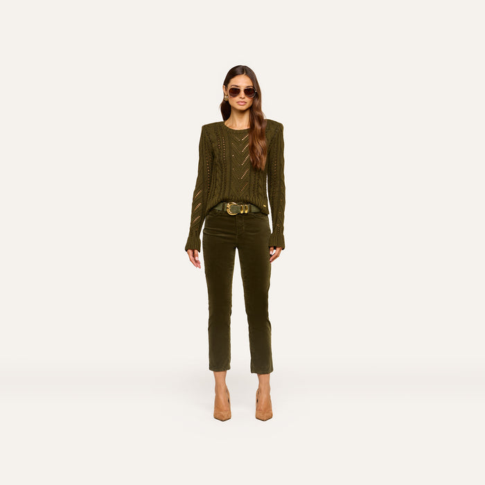 A woman wears the Celestia Velvet Slim-Leg Jean in dark green, paired with an olive green sweater, a brown belt, and beige heels. She has long wavy hair, sunglasses, and stands against a plain light background.