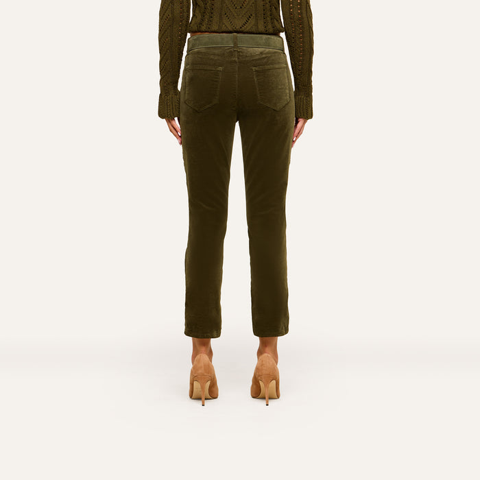 A person stands facing away against a plain background, showcasing the back view of the outfit featuring the Celestia Velvet Slim-Leg Jean in dark green, paired with a matching knit sweater and tan high heels.