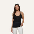 A woman with long brown hair wears the Celia Scoopneck Tank, a sleeveless black featherweight jersey top with a scoop neckline, paired with white pants. She stands against a plain light background and smiles gently at the camera.