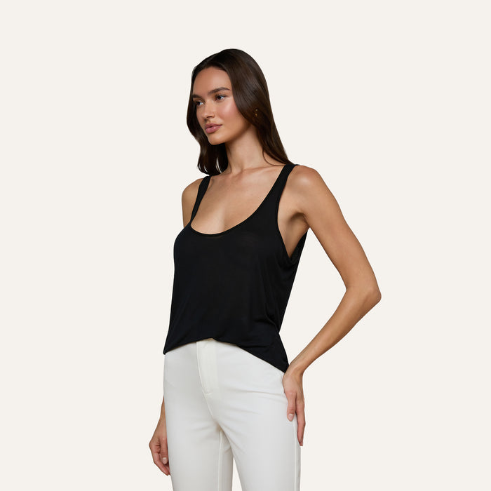 A woman wearing the Celia Scoopneck Tank in black with white pants stands against a plain light background, looking to the side with one hand on her hip.
