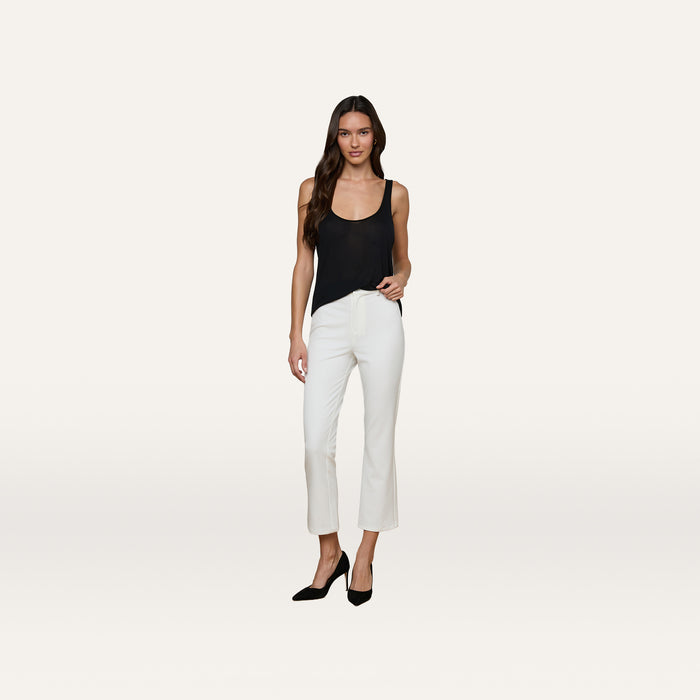 A woman stands against a plain white background wearing the Celia Scoopneck Tank in black, paired with white cropped pants and black heels. She has long brown hair and adopts a relaxed pose.